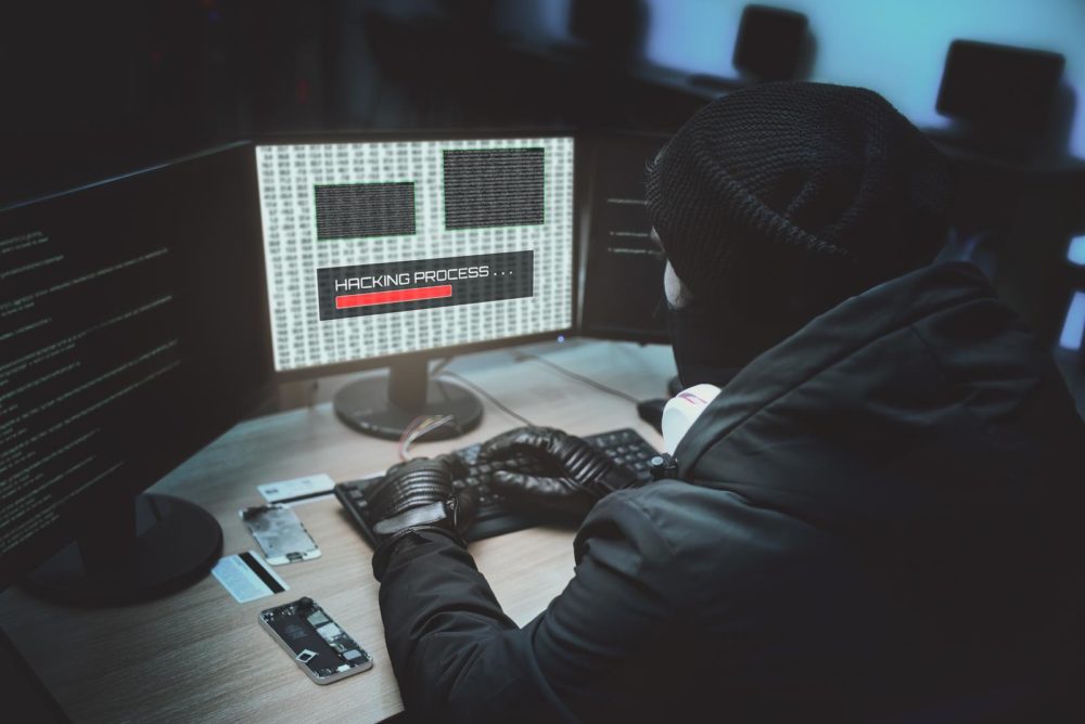 Shot from the Back to Hooded Hacker Breaking into Corporate Data Servers from His Underground Hideout. Place Has Dark Atmosphere, Multiple Displays