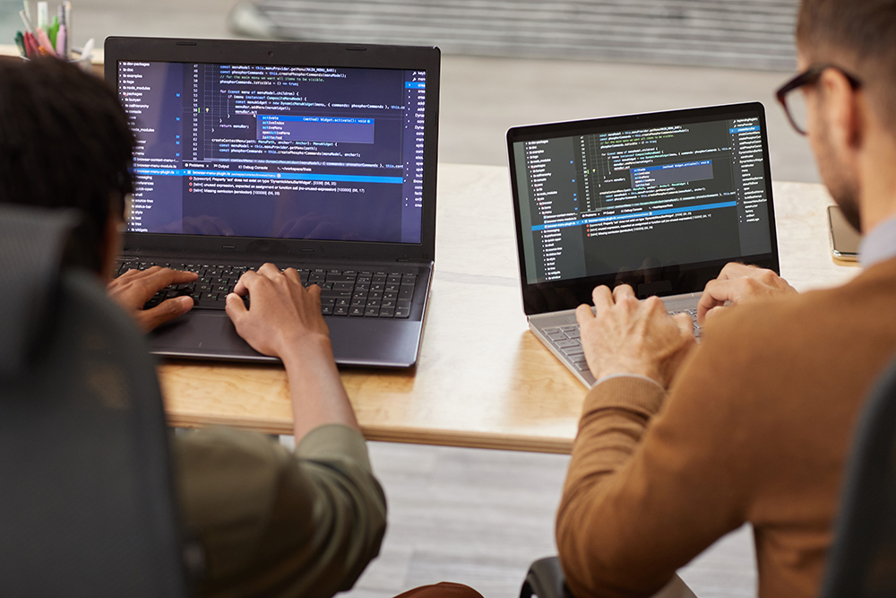 Rear view of two graphic designers sitting at the table and typing on their computers they developing new software