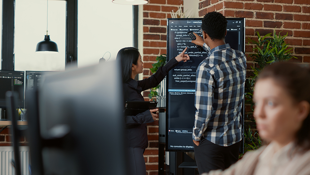 System developers analyzing code on wall screen tv looking for errors comparing results on digital tablet next to team of coders. Programmers working together in it startup company.