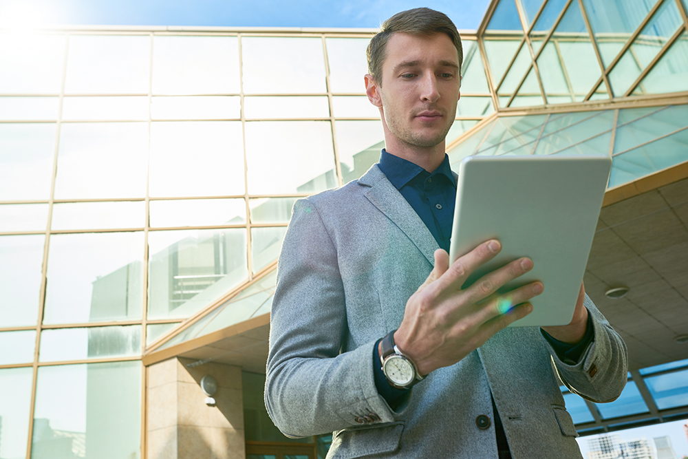 Portrait of handsome man using digital tablet standing outside of modern office building, copy space