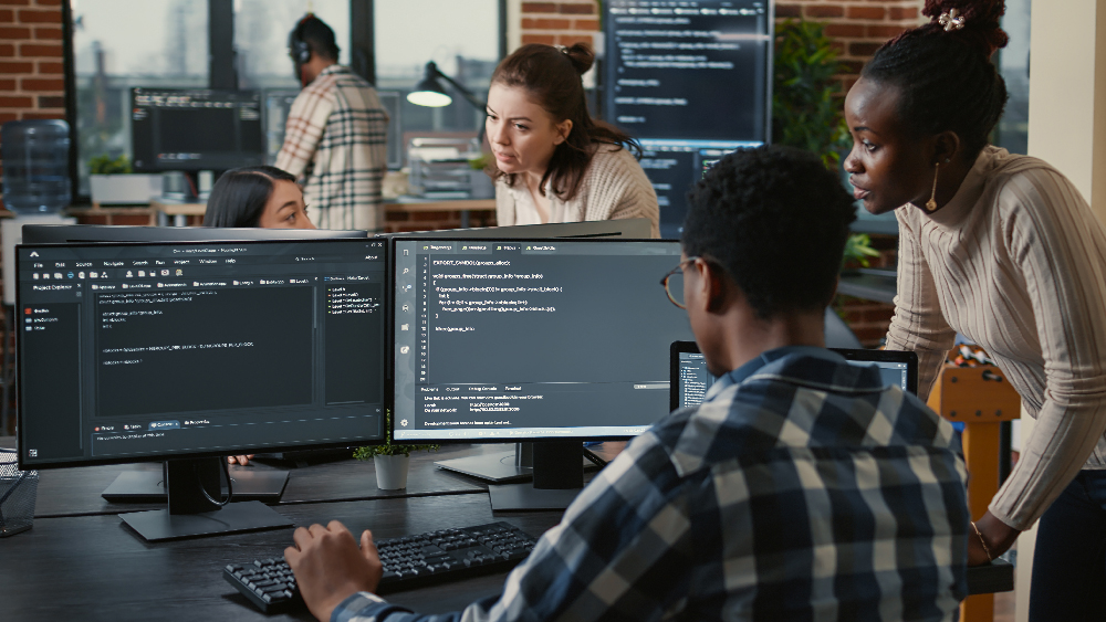 Mixed team of artificial intelligence cloud programers talking about programming in front of multiple screens compiling code. Programmers doing teamwork looking at machine learning algorithms.