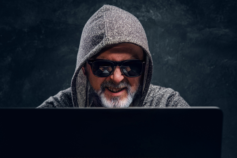 Portrait of old man hacker dressed in sunglasses and hoody using laptop against dark background.