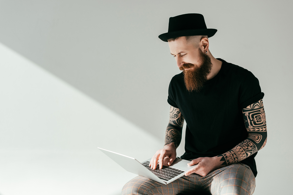 handsome bearded tattooed Black hat hacker looking at laptop 