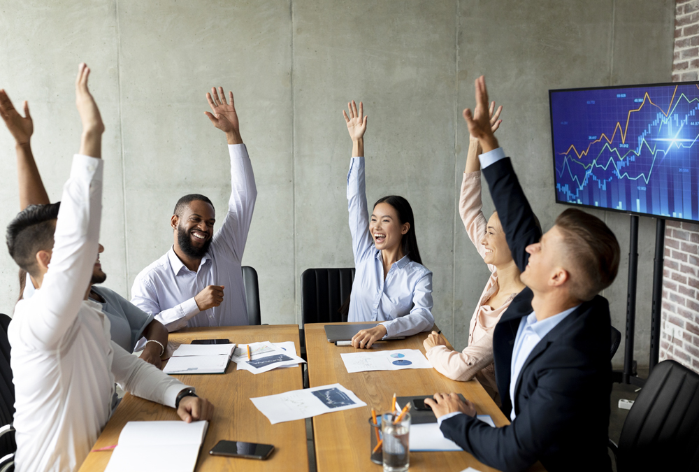 Cybersecurity Training. Group Of Happy Colleagues Raising Hands At Meeting In Office, Multiethnic Coworkers Celebrating Successful Teamwork And Company Growth While Sitting At Desk In Boardroom