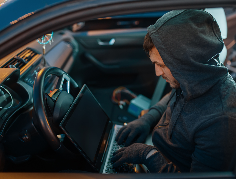 Professional car thief with laptop hacking security system, criminal lifestyle. Hooded male robber opening vehicle on parking. Auto robbery, automobile crime