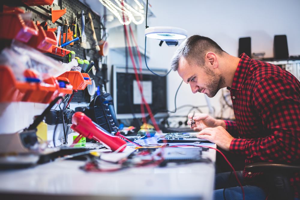 Side view of young concentrated guy repairing laptop and sitting at table between different gadgets and tools