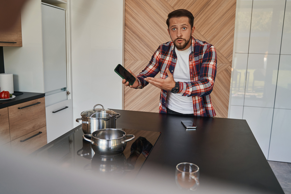 Indignant bearded man in casual clothes with mobile phone in hand standing in the modern kitchen