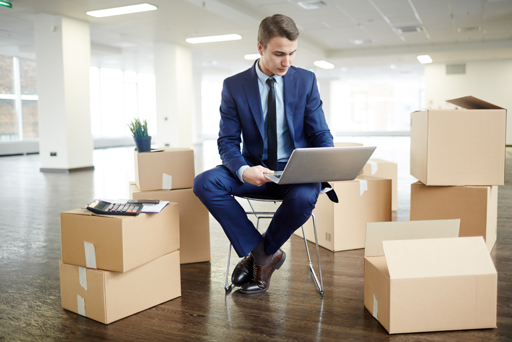 Young employee in elegant suit sitting on chair among unpacked boxes in new office and browsing in the net