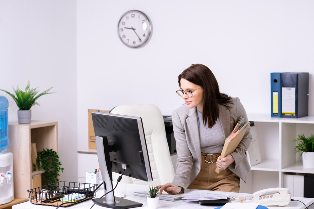 Brunette businesswoman in jacket standing at desk and shutting down computer while leaving office