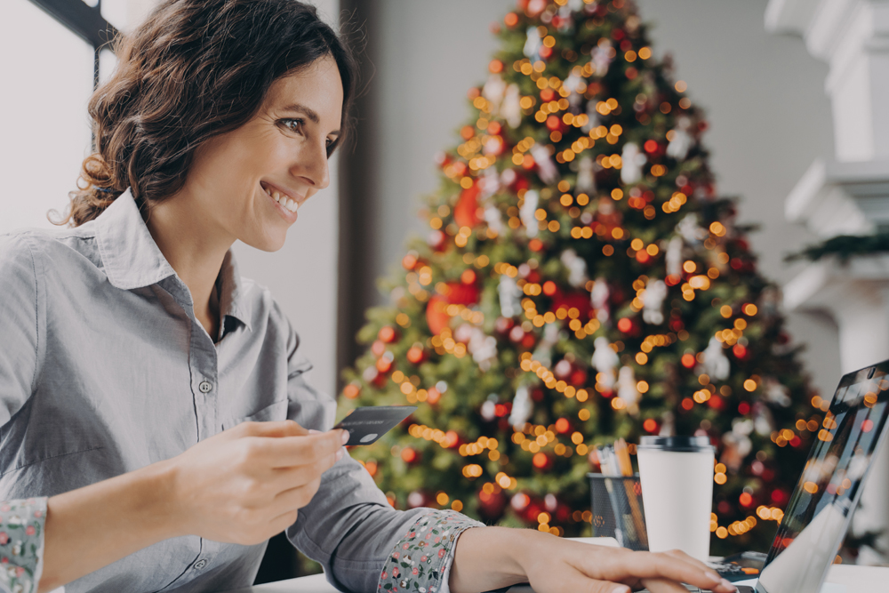 Happy young woman doing online shopping on laptop computer at home during Winter holidays sales, smiling hispanic female with credit card making payment while sitting at table near xmas tree