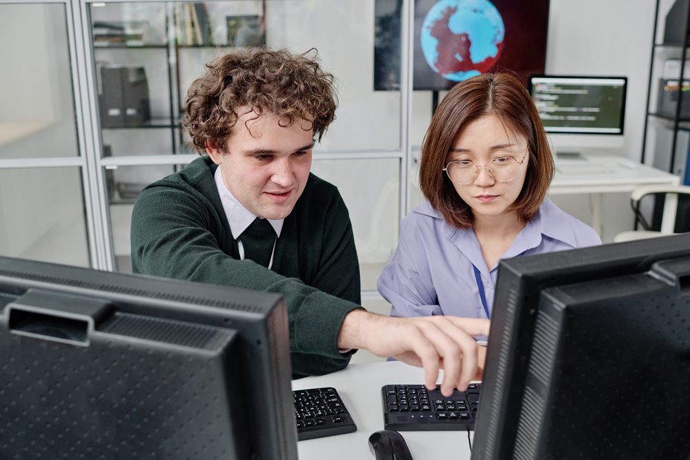 Two young security professionals are working together, they are sitting at table with computer monitors