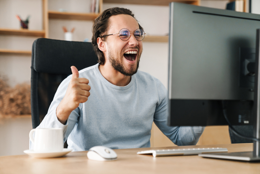 Image of excited programmer man showing thumb up while working with computer in office