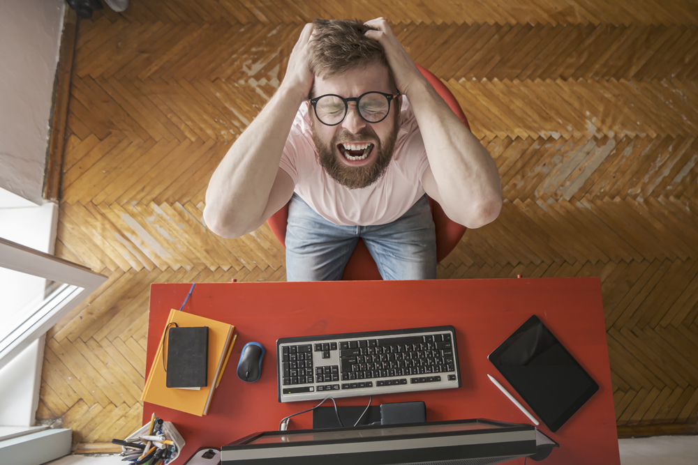 Man experiences stress remotely working at home on the computer, he grabs hands on head and screams loudly. Concept of self-isolation during a pandemic. Top view..