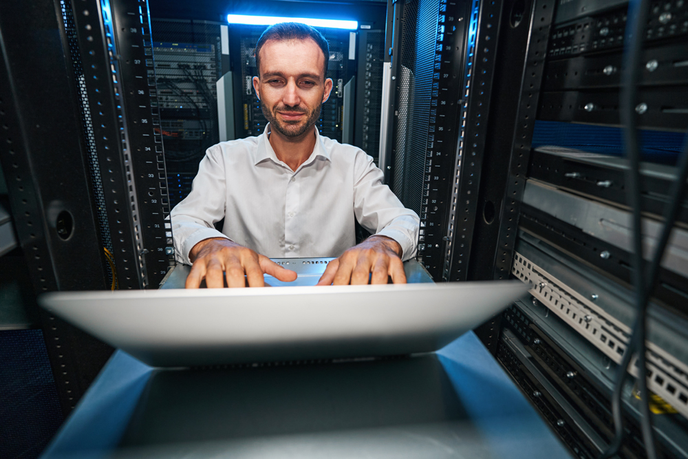 Caucasian male IT technician working on laptop inside server room of data retention center
