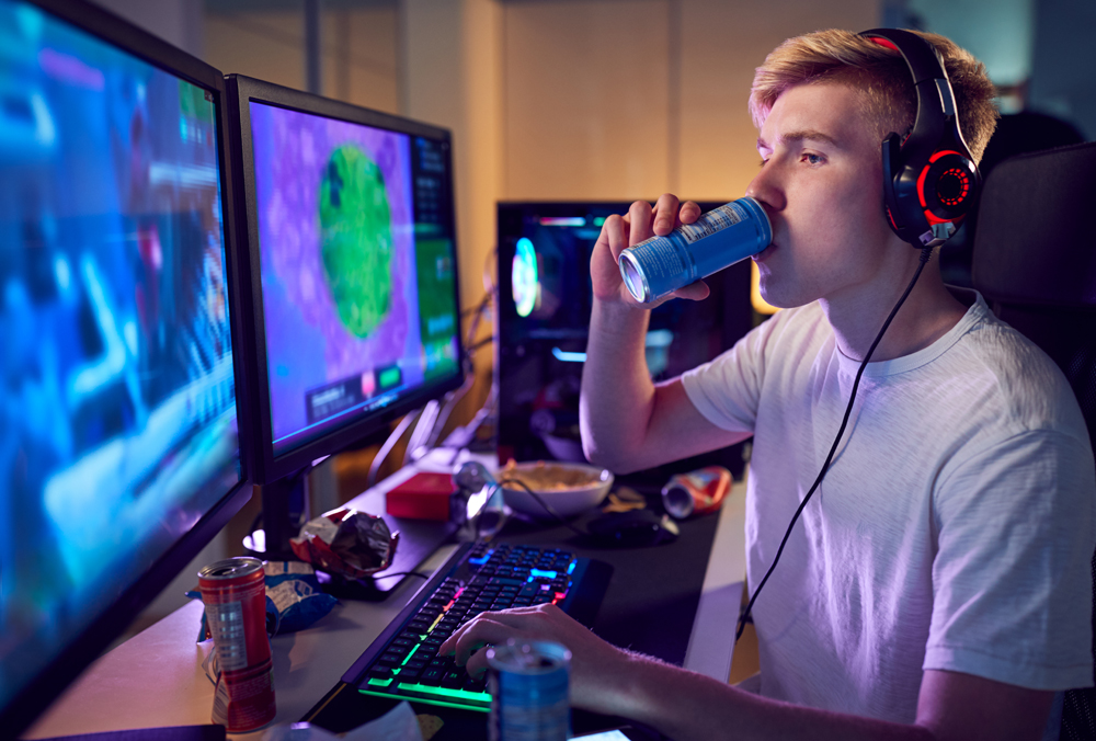 Teenage Boy Drinking Caffeine Energy Drink Gaming At Home Using Dual Computer Screens At Night Thinking ot Himself "is the GPU shortage over?"