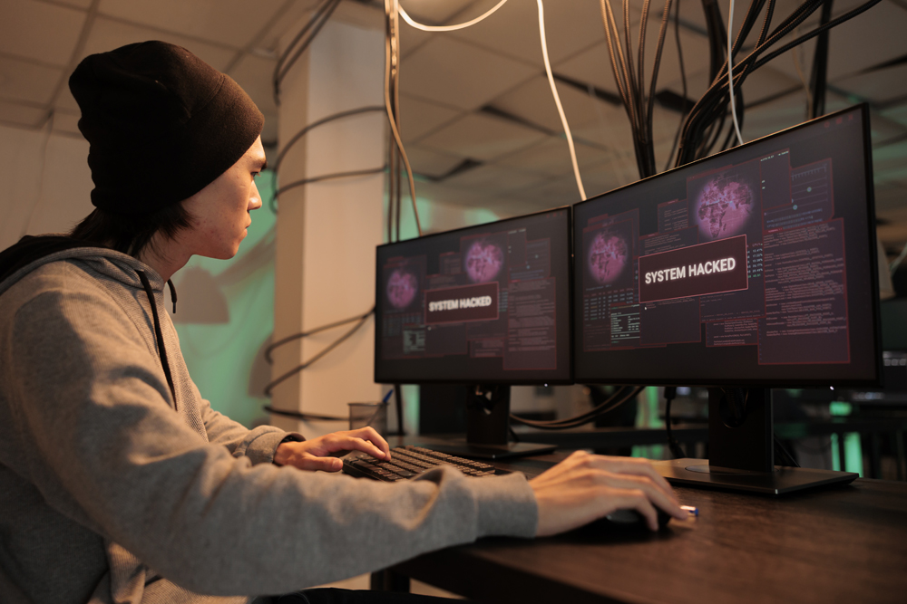 Young Hacker that is sitting in front of multiple computer screens that say System Hacked